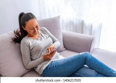 Portrait of smiling Asian woman using smartphone at home sitting on comfortable sofa and reading text messages. Smiling asian woman on couch using phone at home in the living room 