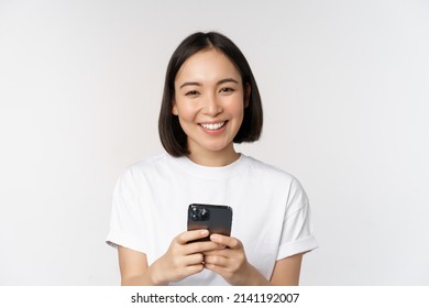 Portrait Of Smiling Asian Woman Using Mobile Phone, Chatting, Texting Message, Standing In Tshirt Over White Background