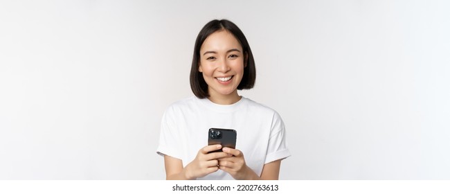 Portrait Of Smiling Asian Woman Using Mobile Phone, Chatting, Texting Message, Standing In Tshirt Over White Background