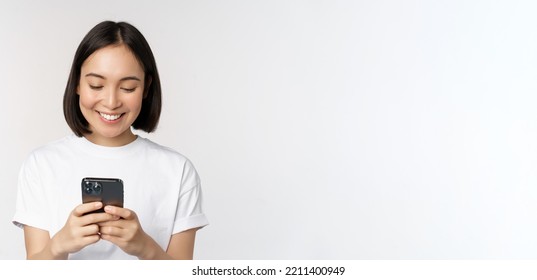 Portrait Of Smiling Asian Woman Using Mobile Phone, Chatting, Texting Message, Standing In Tshirt Over White Background