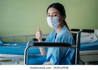 Portrait Of A Smiling Asian Young Female Patient With Surgical Mask Sitting On Wheelchair In Hospital Room Showing Thumbs Up Gesture And Sign To Camera