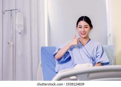 Portrait Of A Smiling Asian Young Female Patient Sitting On Hospital Bed Room Showing Thumbs Up Gesture And Sign To Camera On Recovery From Sickness