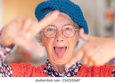 Portrait Of Smiling Attractive Older Woman Wearing Cap And Glasses Gesturing Camera Frame With Hands Looking At Camera With Tongue Sticking Out Of Mouth. Funny Granny Makes Faces