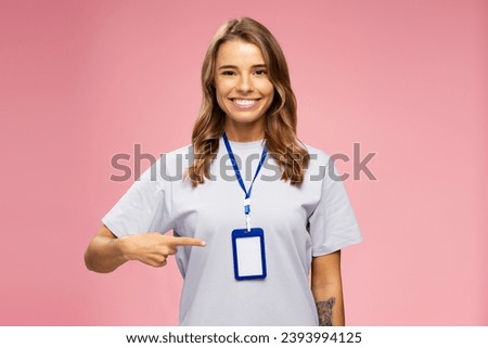 Portrait smiling attractive woman, seller consultant, manager wearing t shirt, pointing on blank badge looking  at camera isolated on pink background. Name card, Mockup