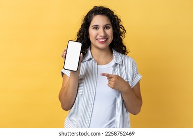 Portrait of smiling attractive young adult woman with dark wavy hair holding pointing at cell phone with empty display, copy space for advertisement. Indoor studio shot isolated on yellow background. - Shutterstock ID 2258752469