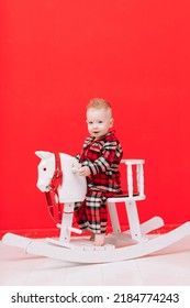 Portrait Of Smiling Baby Boy Sits On Rocking Horse On The Red Background. Happy Little Child In Red Checked Pyjamas Playing With Rocking Chair Children's Toy Vintage. Childcare, Childhood. Copy Space.