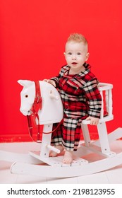 Portrait Of Smiling Baby Boy Sits On Rocking Horse On The Red Background. Happy Little Child In Red Checked Pyjamas Playing With Rocking Chair Children's Toy Vintage. Childcare, Childhood. Copy Space.