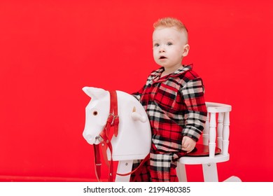 Portrait Of Smiling Baby Boy Sits On Rocking Horse On The Red Background. Happy Little Child In Red Checked Pyjamas Playing With Rocking Chair Children's Toy Vintage. Childcare, Childhood. Copy Space.