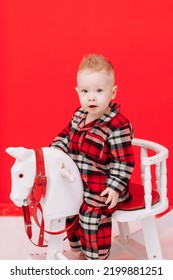 Portrait Of Smiling Baby Boy Sits On Rocking Horse On The Red Background. Happy Little Child In Red Checked Pyjamas Playing With Rocking Chair Children's Toy Vintage. Childcare, Childhood. Copy Space.