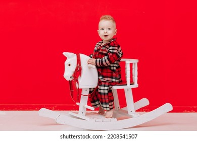 Portrait Of Smiling Baby Boy Sits On Rocking Horse On The Red Background. Happy Little Child In Red Checked Pyjamas Playing With Rocking Chair Children's Toy Vintage. Childcare, Childhood. Copy Space.