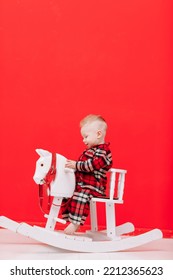 Portrait Of Smiling Baby Boy Sits On Rocking Horse On The Red Background. Happy Little Child In Red Checked Pyjamas Playing With Rocking Chair Children's Toy Vintage. Childcare, Childhood. Copy Space.