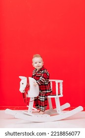 Portrait Of Smiling Baby Boy Sits On Rocking Horse On The Red Background. Happy Little Child In Red Checked Pyjamas Playing With Rocking Chair Children's Toy Vintage. Childcare, Childhood. Copy Space.