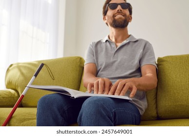 Portrait of a smiling bearded blind man in black eyeglasses sitting on sofa in the living room at home with stick cane and reading tactile braille book. Blindness and education concept. - Powered by Shutterstock