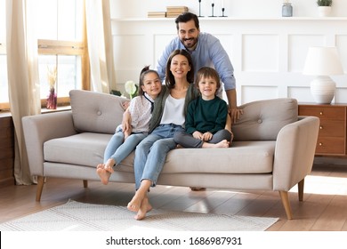 Portrait of smiling bearded father standing near couch with sitting happy wife and little children. Joyful affectionate family of four posing for photo, looking at camera, good relations concept. - Powered by Shutterstock