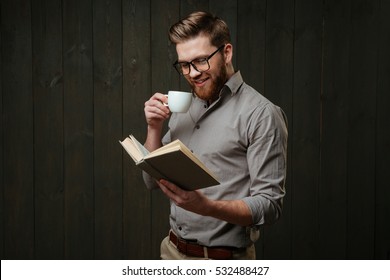 Portrait Of A Smiling Bearded Man In Eyeglasses Reading Book And Drinking Cup Of Coffee Isolated On A Black Wooden Background