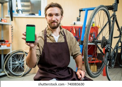 Portrait of smiling bearded mechanic sitting on haunches and showing smartphone with blank screen to camera, interior of modern bicycle repair shop on background - Powered by Shutterstock