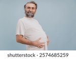 Portrait of a smiling bearded senior man holding his big belly with hands on plain studio background