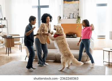 Portrait of smiling beautiful African American family, mother, daughter and son playing dancing with golden retriever at home in living room. Children having fun. Happy childhood concept - Powered by Shutterstock