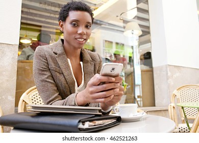 Portrait Of Smiling Beautiful African American Business Woman Sitting In A Classic City Coffee Shop Using A Smart Phone, Looking, Exterior. Professional Black Woman, Technology Lifestyle Outdoors.