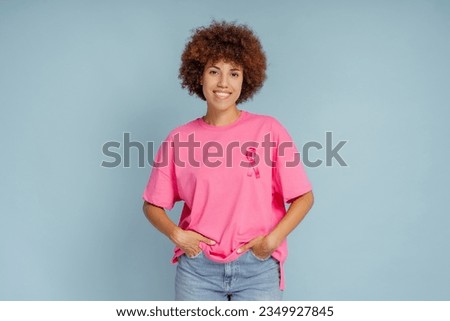 Portrait of smiling beautiful African American woman wearing t shirts with pink ribbon isolated on blue background. Health care, support, prevention. Breast cancer awareness month concept