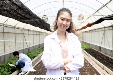 Portrait of smiling beautiful Asian botanist scientist woman in white lab coat standing with arms crossed. Female biological researcher work on experimental plant in greenhouses. Agricultural Science - Powered by Shutterstock