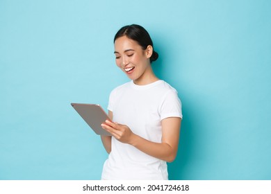Portrait of smiling beautiful asian girl, female 3d artist or freelancer using digital tablet and looking pleased at device screen, shopping online or browsing over light blue background