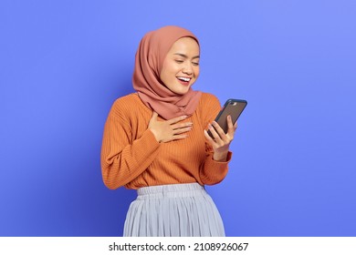 Portrait of smiling beautiful Asian woman in brown sweater and hijab holding on mobile phone with hand on chest isolated over purple background. People islam religious concept - Shutterstock ID 2108926067