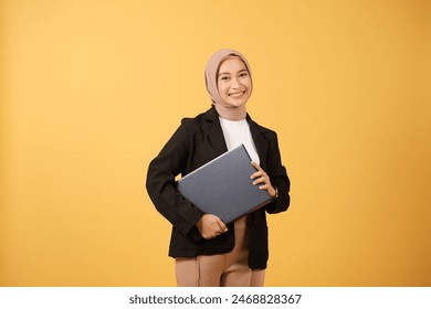 Portrait of smiling beautiful Asian woman in casual shirt and hijab holding a laptop, showing thumb up isolated over yellow background. People religious lifestyle concept - Powered by Shutterstock