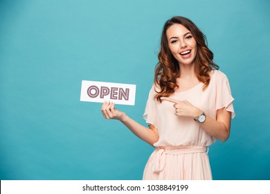 Portrait of a smiling beautiful girl wearing dress and pointing finger at an open sign isolated over blue background