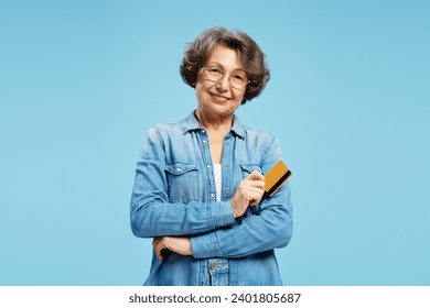 Portrait of smiling beautiful gray haired senior woman holding credit card, isolated on blue background. Concept of banking, advertisement