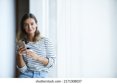 Portrait Of Smiling Beautiful Hipster Designer In Stripped Sweater Drinking Takeout Coffee And Using Smartphone