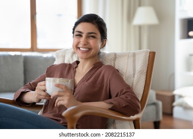 Portrait of smiling beautiful indian ethnicity woman resting in cozy armchair with cup of coffee or tea in hands, enjoying carefree lazy relaxed morning weekend time lone in modern living room. - Shutterstock ID 2082215137