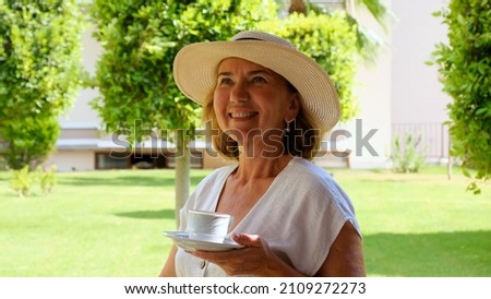 Portrait of smiling beautiful mature female relaxing in her garden with hot drink coffee in morning on a sunny day. Concept of waking up in the morning, planning things for the day, daydreaming