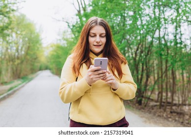 A portrait of a smiling beautiful redhead woman texting sms with her phone on nature background. Happy woman is using a smartphone in park outdoors, spring time. Traveler - Shutterstock ID 2157319041