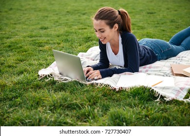 Portrait Of A Smiling Beautiful Student Girl Doing Homework On Laptop Computer While Lying On The Grass At Park