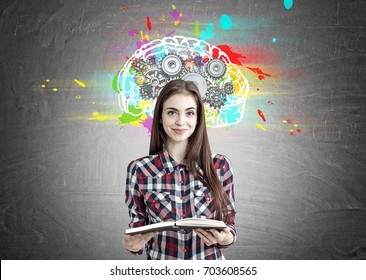 Portrait of a smiling beautiful teenage girl in a checkered shirt holding an open book and standing near a blackboard with a colorful brain sketch and gears on top of it