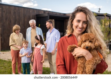 Portrait Of Smiling Beautiful Woman Holding Labradoodle Dog, Her Family In Standing In Background