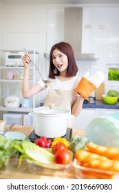 Portrait Of Smiling Beautiful Young Asian Woman Cooking Soup And Looking At Camera In Light White Interior Style Kitchen