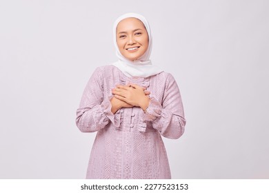 Portrait of smiling beautiful young Asian Muslim woman wearing hijab and purple dress holding hands on chest and greeting welcoming Ramadan isolated on white studio background
