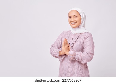 Portrait of smiling beautiful young Asian Muslim woman wearing hijab and purple dress standing with gesturing Eid Mubarak greeting and welcoming Ramadan isolated on white studio background