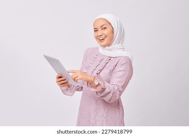 Portrait of smiling beautiful young Asian Muslim woman wearing hijab and purple dress holding digital tablet isolated on white studio background - Powered by Shutterstock