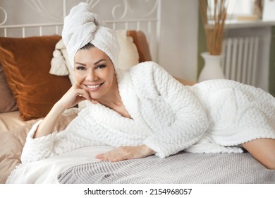 Portrait of smiling beautiful young woman lies on bed in a cozy bedroom in bathrobe and towel on her head after spa at hotel