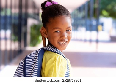 Portrait of smiling biracial schoolgirl with school bag, turning to camera outside school. Education, inclusivity, childhood, elementary school and learning concept. - Powered by Shutterstock