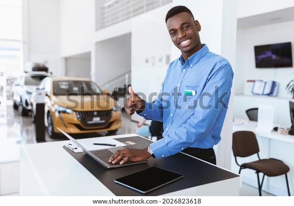 Portrait of\
smiling black car salesman showing thumb up gesture while using\
laptop at automobile dealership. Cheerful Afro manager posing at\
workplace, looking at camera in auto\
showroom