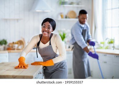 Portrait Of Smiling Black Lady In Apron And Rubber Gloves Wiping Kitchen Table, Her Boyfriend Washing Floor On Background, Blank Space. Positive African American Couple Cleaning Their House