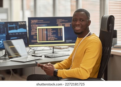 Portrait of smiling black man looking at camera while sitting at workplace in software development company against computer screens with code