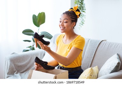 Portrait of smiling black woman received package, unpacking cardboard box with shoes, holding and looking at high heels, sitting on the sofa at home. Female buyer satisfied with online shopping