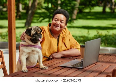 Portrait Of Smiling Black Woman Using Laptop In Outdoor Cafe With Cute Pug Dog Scene Lit By Sunlight