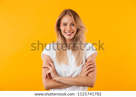 Portrait of a smiling blonde young woman standing with arms folded isolated over yellow background