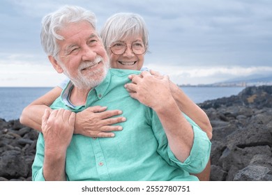 Portrait of smiling bonding senior couple embracing sitting outdoors on the seaside expressing love and affection. Cloudy sky in background and horizon over water - Powered by Shutterstock